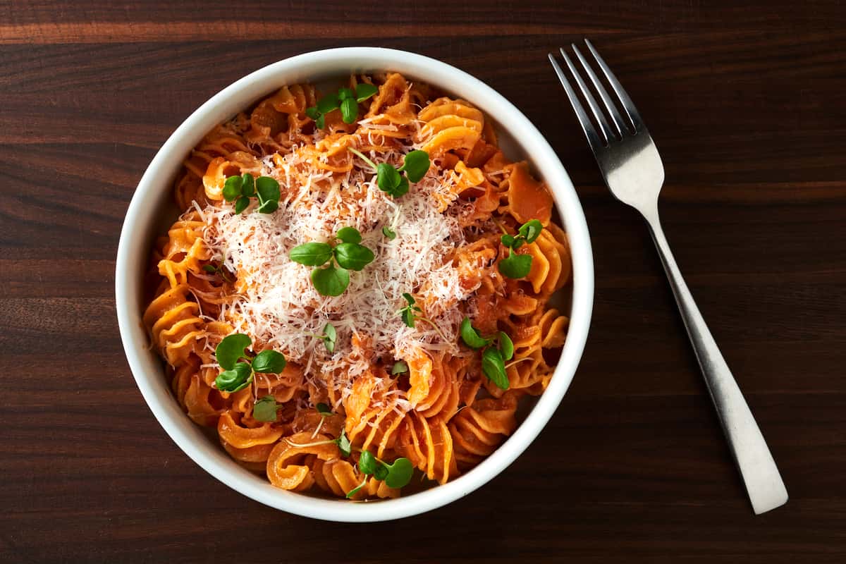Overhead view of a bowl of radiatore pasta tossed in creamy vodka sauce, topped with grated Parmesan cheese and fresh microgreens, served on a dark wood table with a fork beside the bowl.