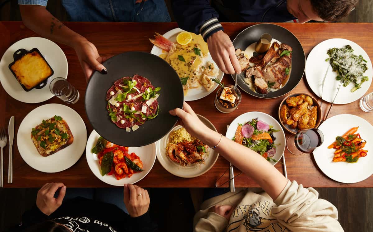 A lively overhead view of a group sharing a colorful Italian feast at Sofia restaurant in New Orleans, featuring a variety of vibrant dishes including seafood, beef carpaccio, roasted meats, salads, and vegetables, all spread across a rich wooden table with hands reaching in to serve and share.