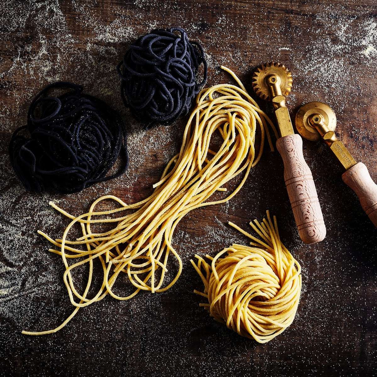 Fresh housemade yellow and black pasta nests arranged on a floured wooden surface beside traditional pasta-cutting tools with wooden handles.