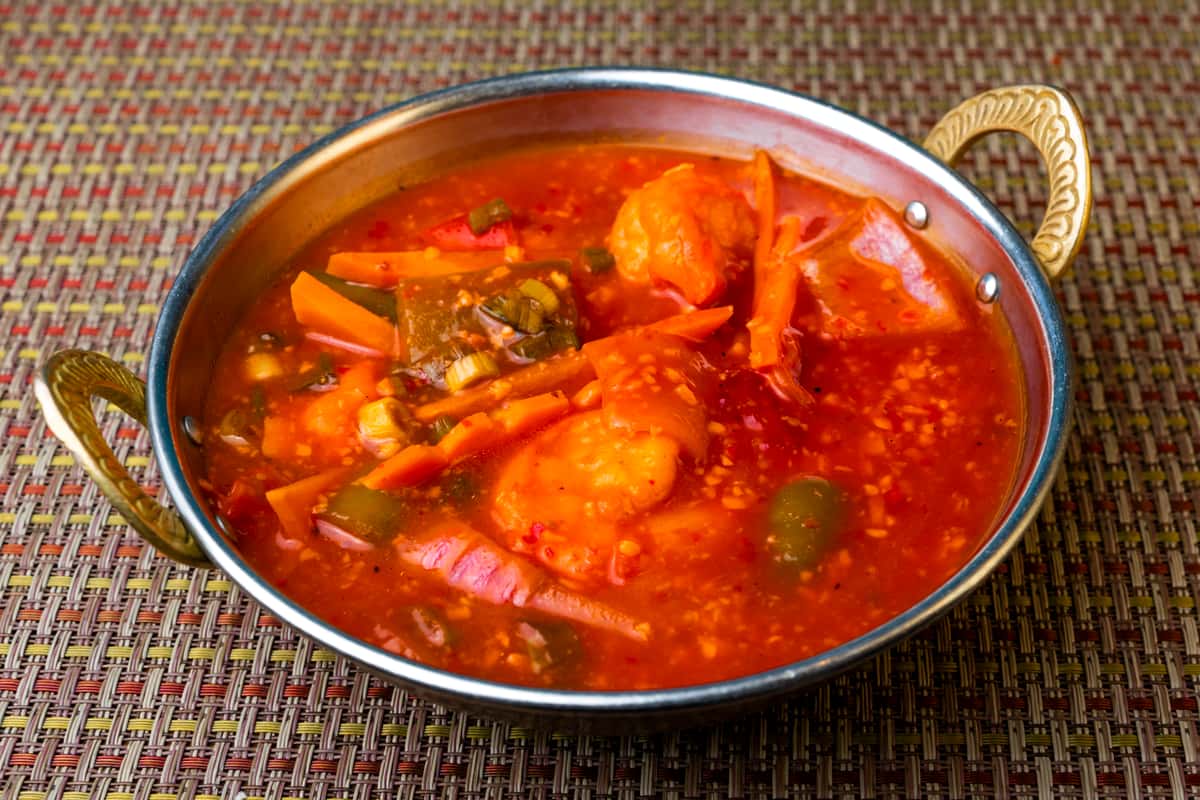 Spicy Indo-Chinese shrimp in a vibrant red chili garlic sauce with carrots, bell peppers, and green onions — bold Asian fusion seafood dish served in a traditional metal bowl.