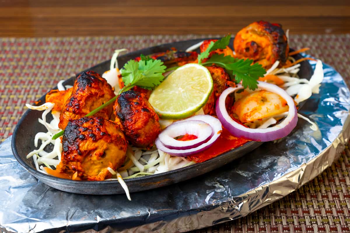 Grilled chicken tikka skewers served on a sizzling platter with shredded cabbage, red onion rings, fresh cilantro, and a lime wedge — classic Indian tandoori appetizer with smoky charred spices.