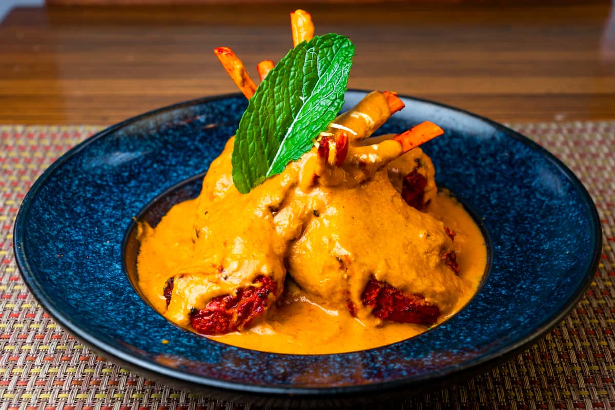 Lamb chops topped with creamy spiced masala sauce and mint garnish, served on a blue plate — rich Indian tandoori-style lamb entrée.