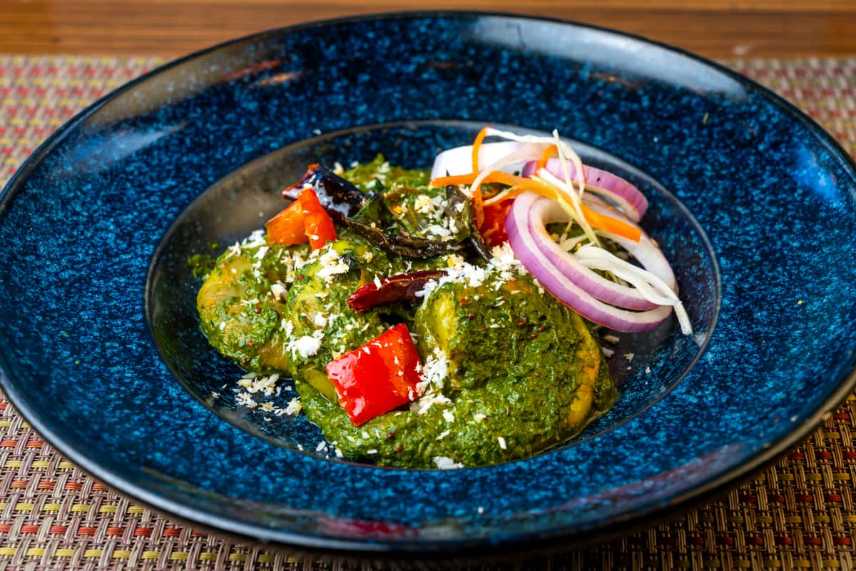 Indian palak paneer served in a deep blue bowl—soft paneer cubes in creamy spinach curry topped with grated cheese, red chili, curry leaves, and fresh onion salad garnish.