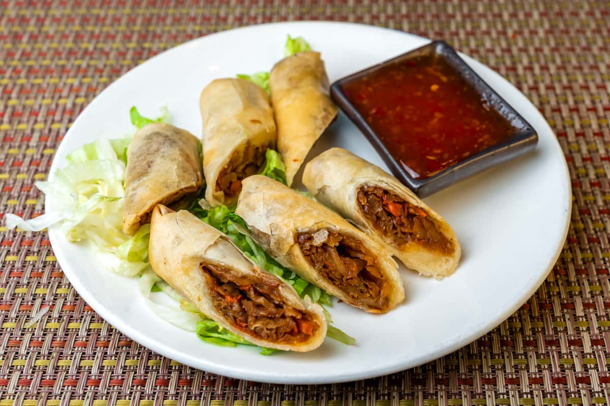 Crispy vegetable spring rolls cut in halves and served with sweet chili dipping sauce on a white plate—golden fried Asian-style appetizer with shredded vegetable filling and lettuce garnish.
