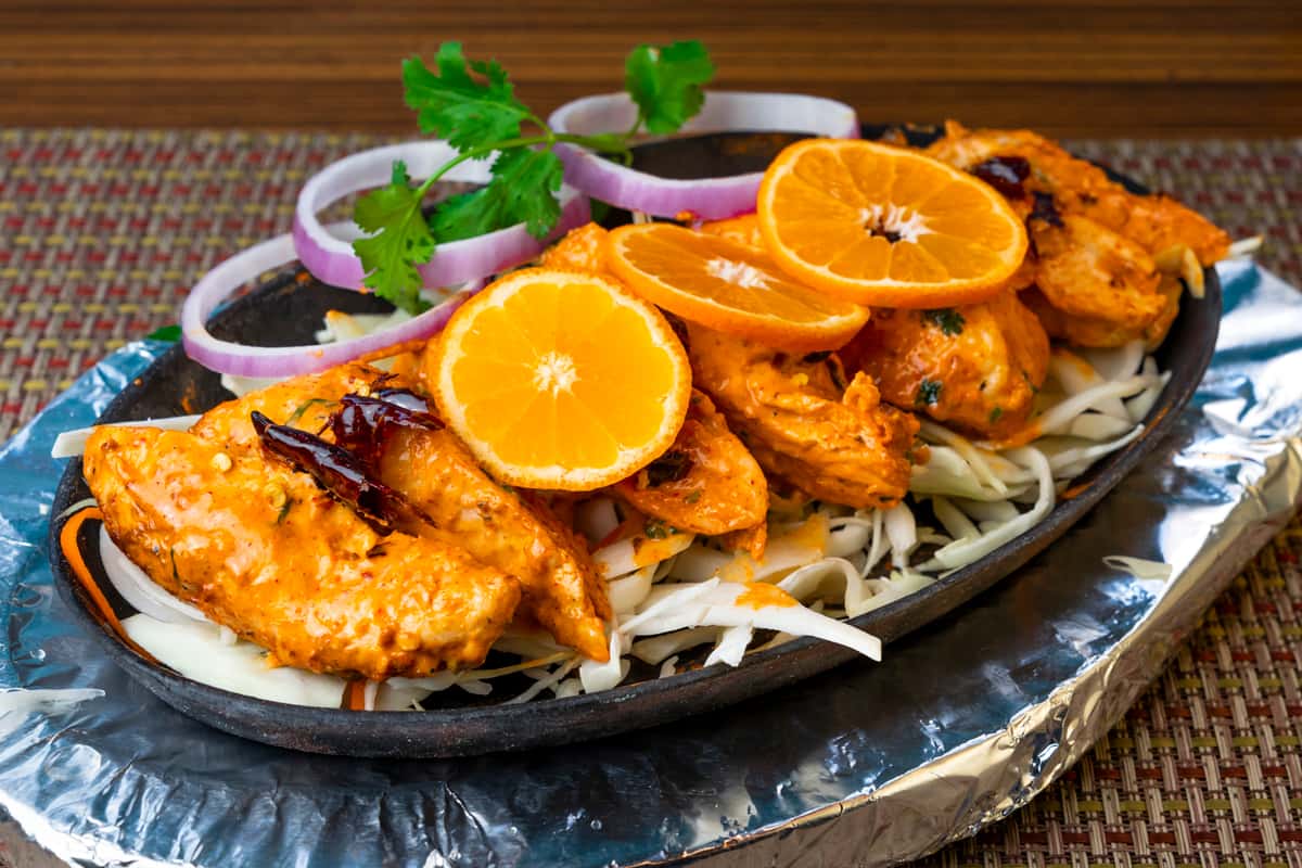 Chicken tikka served on a sizzling platter with shredded cabbage, topped with orange slices, onion rings, and fresh cilantro—flavorful Indian tandoori grilled chicken appetizer.