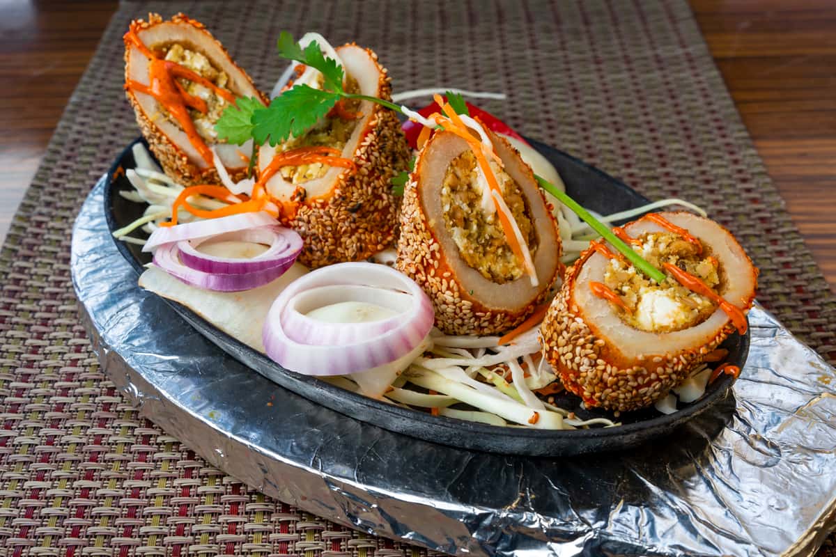 Crispy sesame-crusted stuffed chicken slices served on a sizzling platter with shredded cabbage, red onion rings, and carrot garnish — flavorful Indo-Chinese style appetizer with spiced filling.