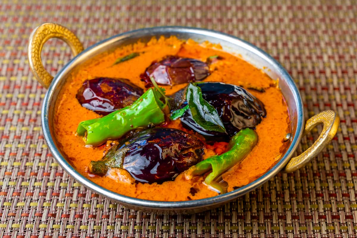 Creamy Indian eggplant curry (baingan) in a rich spiced tomato gravy, topped with whole green chilies and herbs, served in a traditional metal karahi bowl.