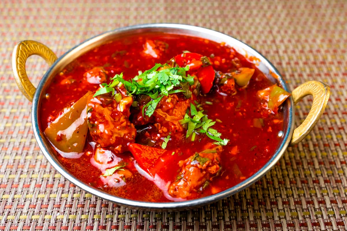 Spicy paneer chili curry in a rich red Indo-Chinese sauce with bell peppers and herbs, served in a traditional Indian metal bowl—vibrant vegetarian paneer dish garnished with fresh cilantro.