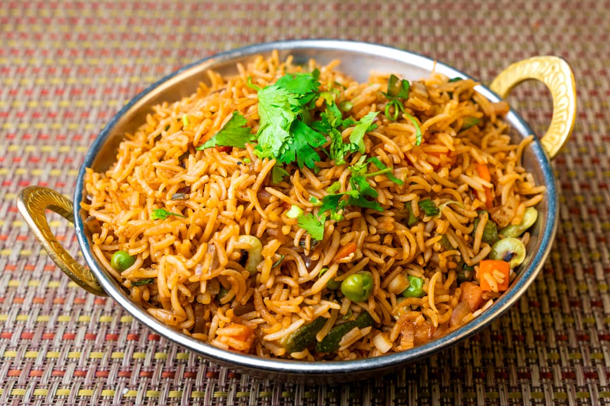 "Vegetable biryani served in a traditional metal bowl, featuring fragrant basmati rice mixed with peas, carrots, and spices, garnished with fresh cilantro on a woven table mat—colorful Indian vegetarian rice dish.