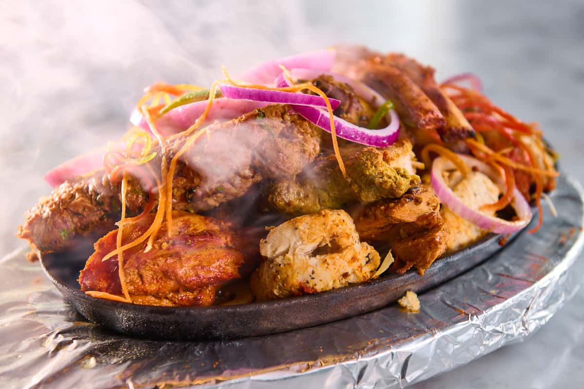 "Sizzling mixed tandoori platter with grilled chicken tikka, seekh kebab, and paneer, topped with red onion rings and shredded carrots—smoky Indian barbecue appetizer served on a hot iron plate.