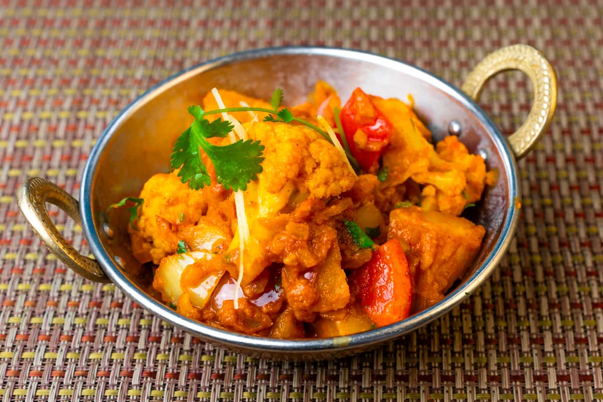 Aloo gobi curry with cauliflower and potatoes cooked in rich Indian spices, served in a traditional metal bowl and garnished with fresh cilantro and ginger.