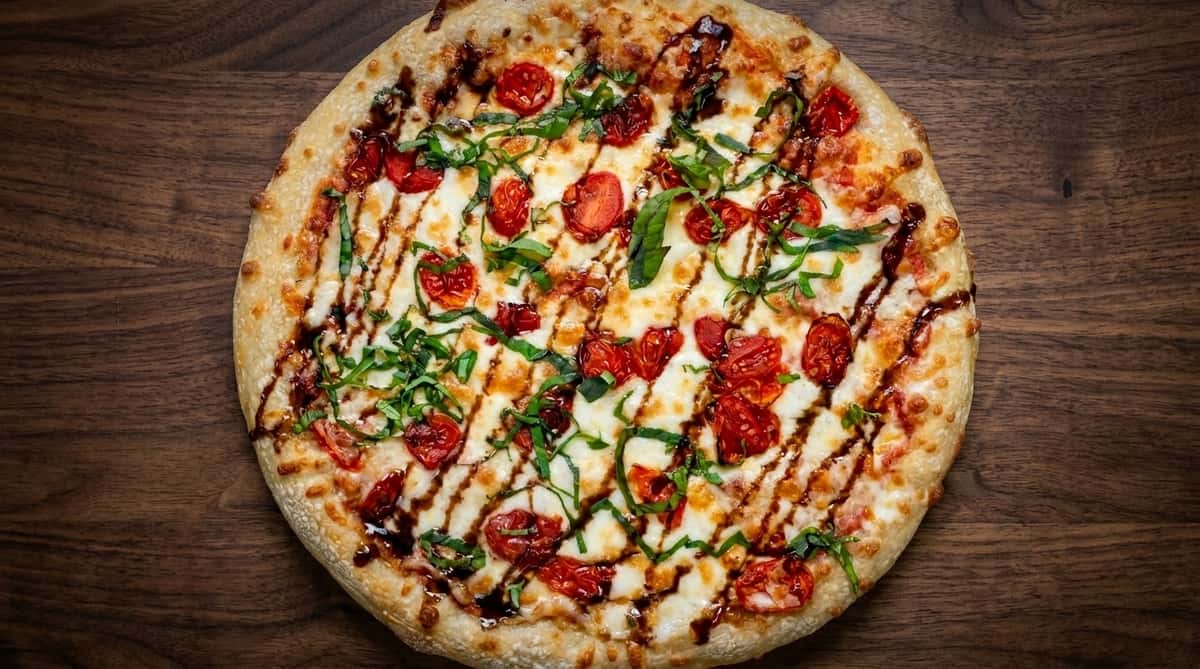 Wood-fired Margherita-style pizza topped with melted mozzarella, roasted cherry tomatoes, fresh basil, and balsamic glaze on a rustic wooden table