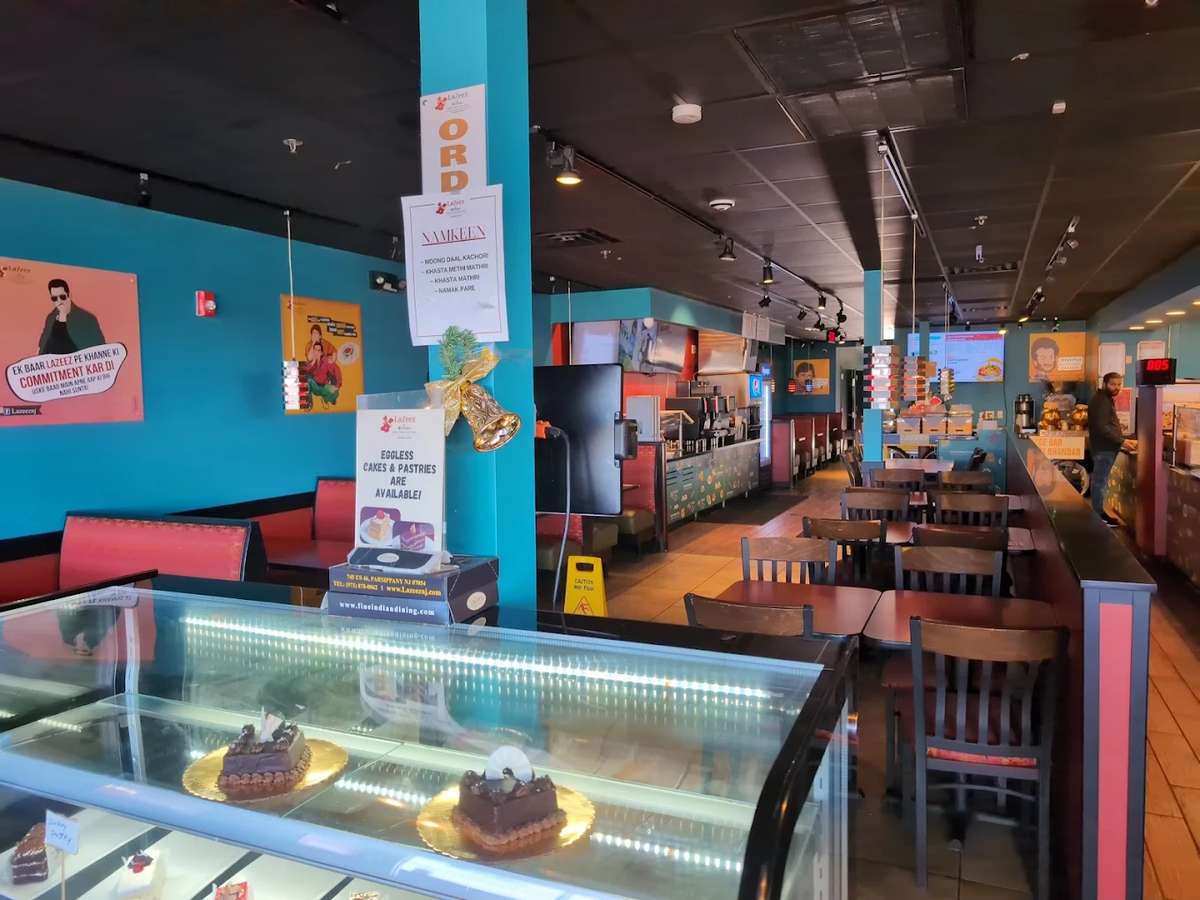 Modern Indian fast-casual restaurant interior featuring teal walls, wooden seating, a dessert display case with chocolate cakes, and a clean, vibrant dining space with counter service and colorful decor.