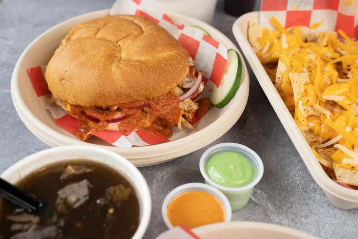 Indian street food–inspired meal featuring a saucy chicken tikka sandwich on a soft bun, loaded masala nachos with melted cheese, tangy chutneys, and a side of spiced soup, served on a casual restaurant table.