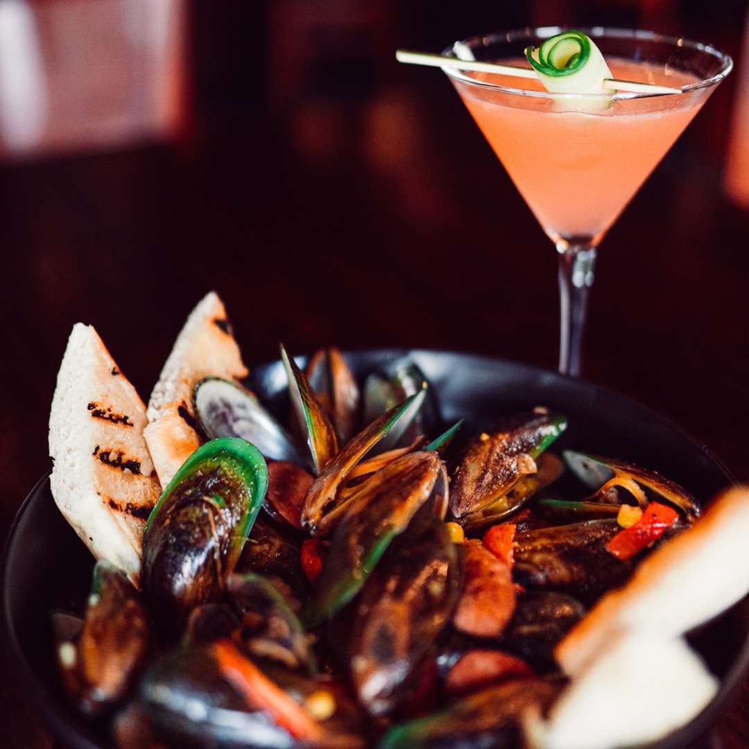 Bowl of steamed mussels with grilled bread and peppers, paired with a pink cocktail garnished with cucumber ribbon.