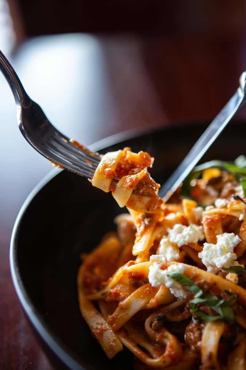 Close-up of tagliatelle pasta in rich tomato sauce with crumbled ricotta cheese and fresh basil, twirled on a fork.