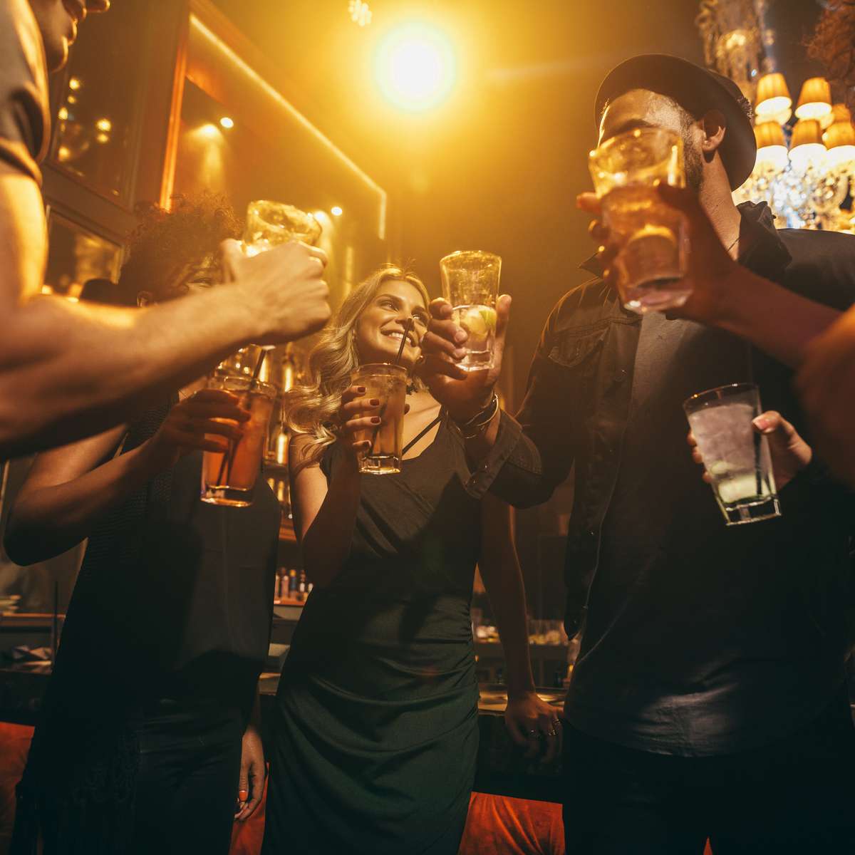 A group of people raising their glasses in a toast, smiling and celebrating in a warm, dimly lit bar atmosphere.