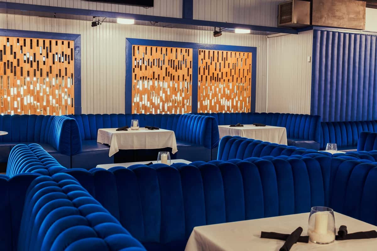 A cozy dining area with plush blue velvet booths and modern wooden panel walls, complemented by clean white tables with candle holders.