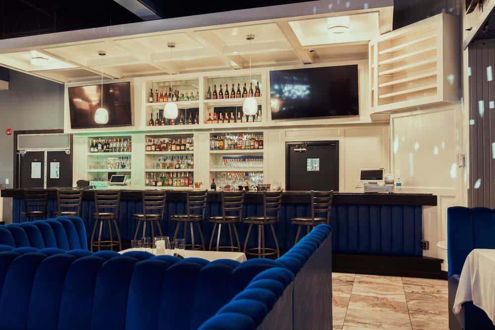 A well-stocked bar with stylish blue seating, backlit shelves holding various liquor bottles, and large flat-screen TVs hanging above the bar area.