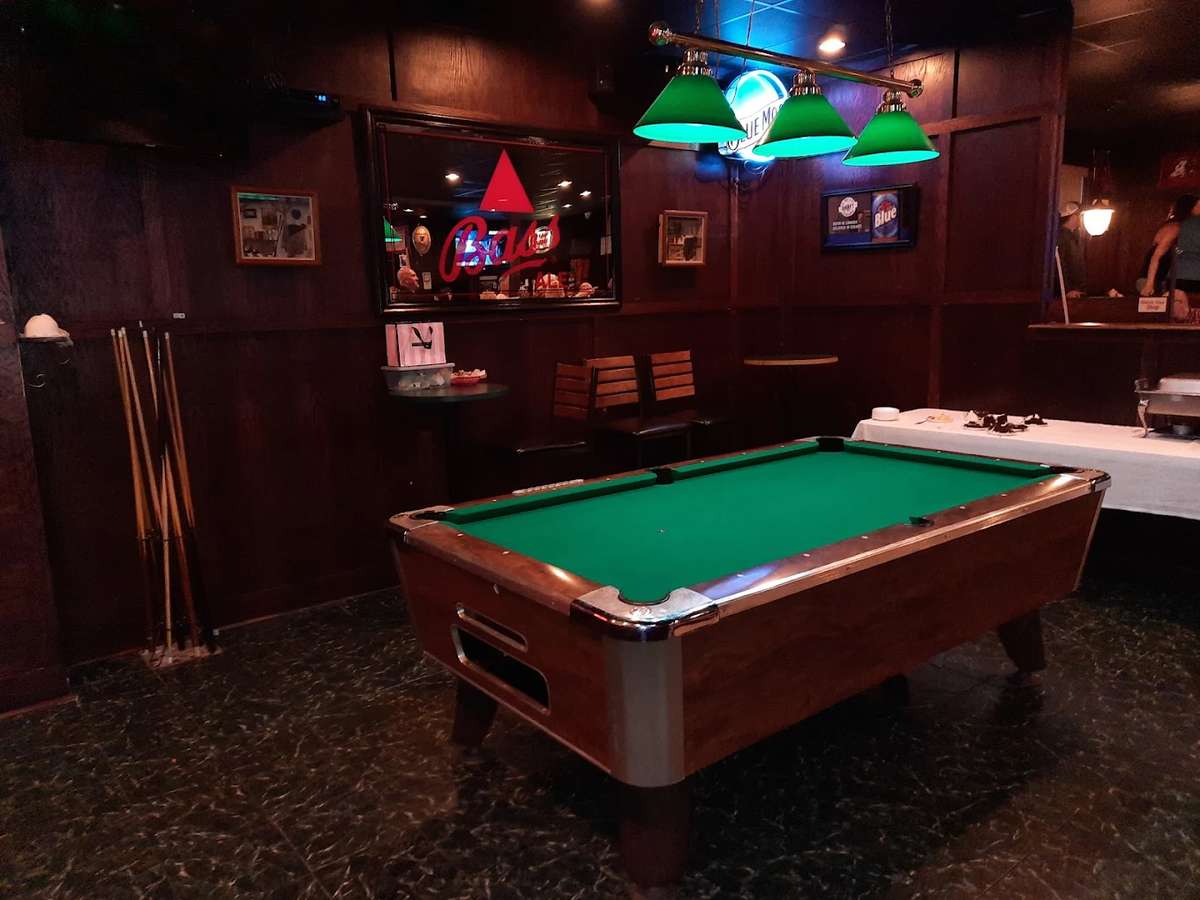 A billiards table sits in a dimly lit room featuring dark wood-paneled walls and a speckled floor. Three green pendant lamps hang directly over the green felt of the pool table, and a small bar area with stools is visible in the background.