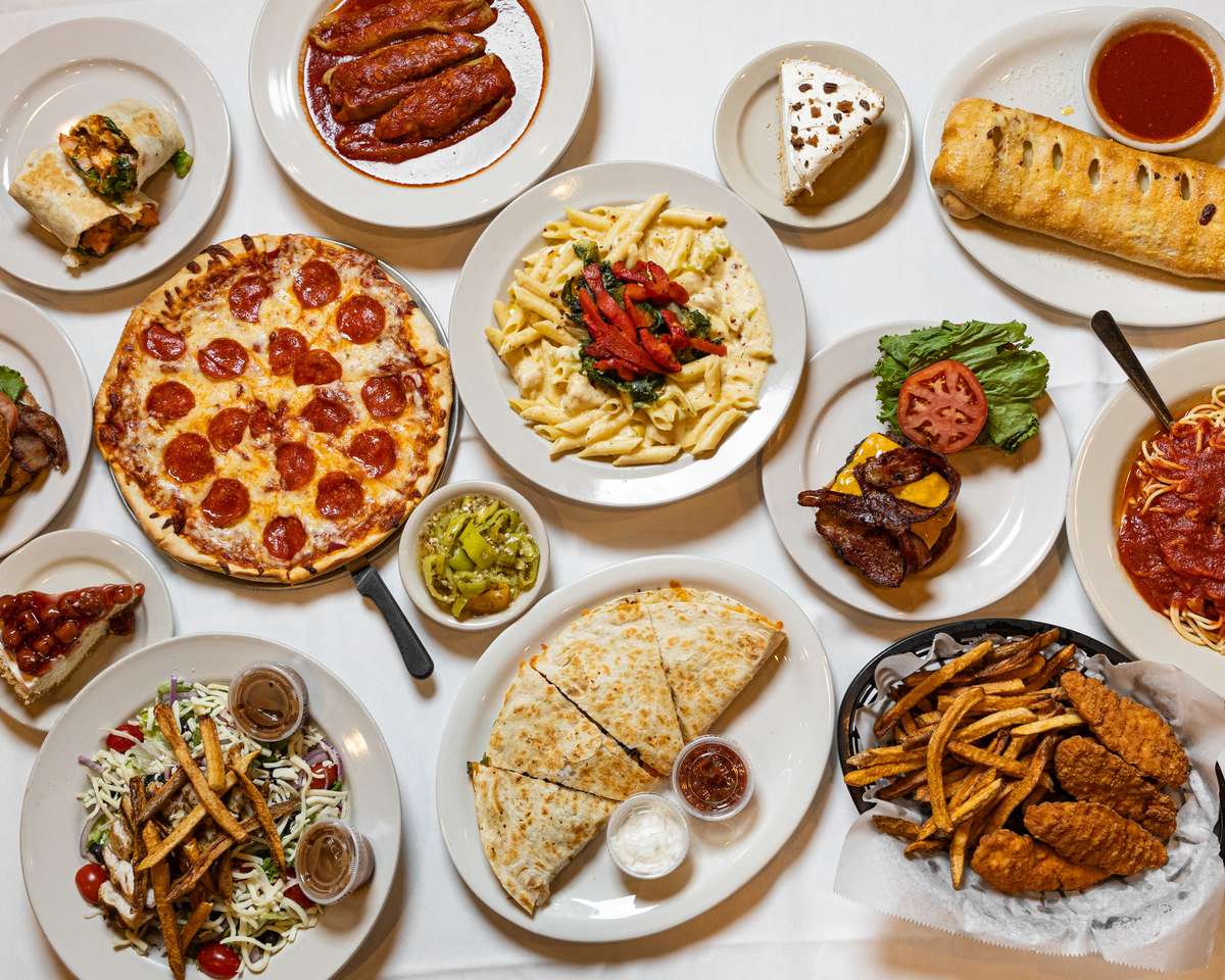 A flat lay of an Italian feast featuring pepperoni pizza, penne pasta, a cheeseburger, quesadillas, chicken tenders, fries, and salad.