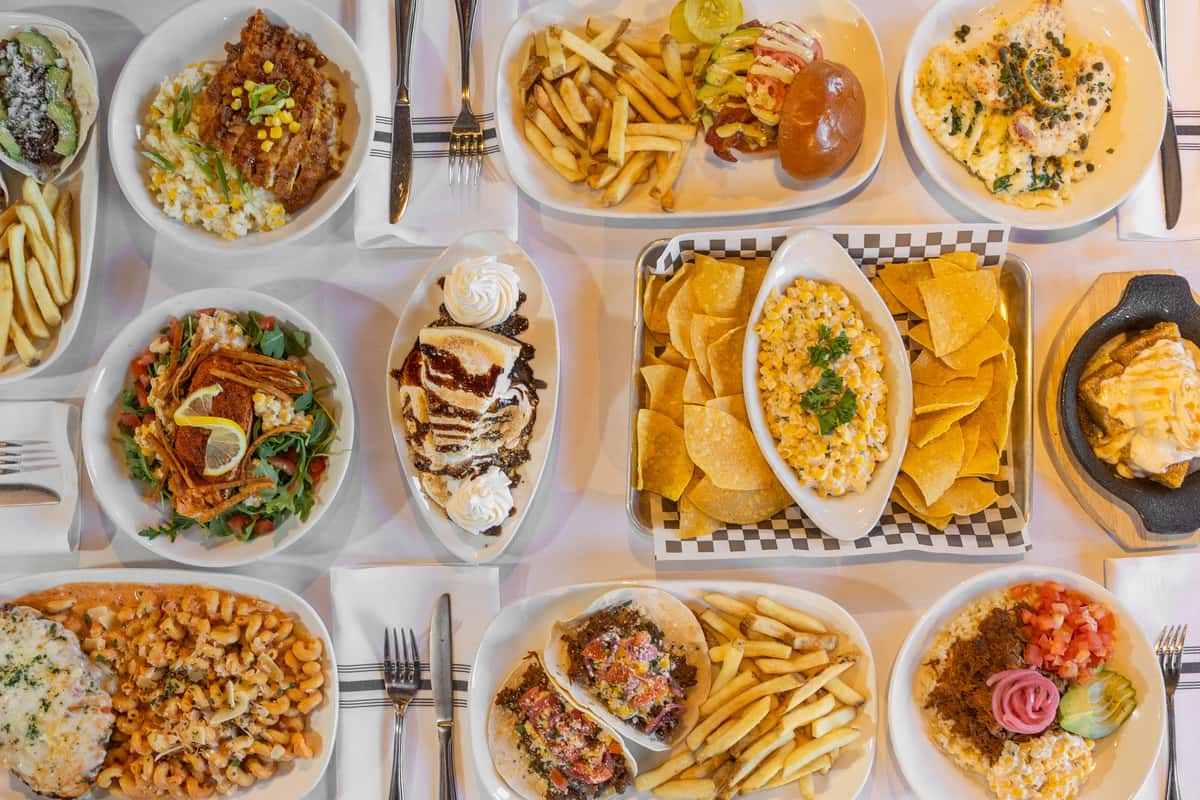 overhead view of assorted restaurant dishes including burgers, pasta, tacos, fries, dips, and desserts on a dining table