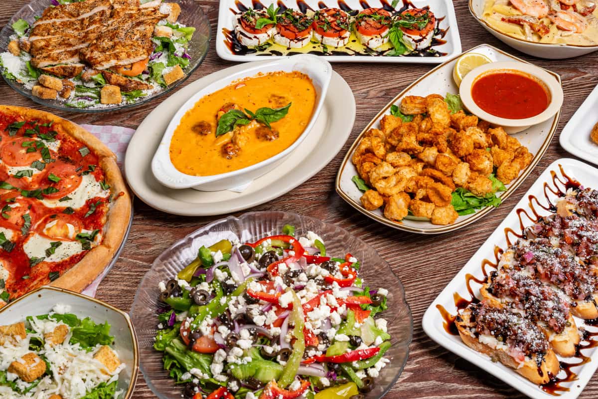 Large Italian food spread featuring pizza, bruschetta, creamy seafood bisque, grilled chicken Caesar salad, and Greek salad.