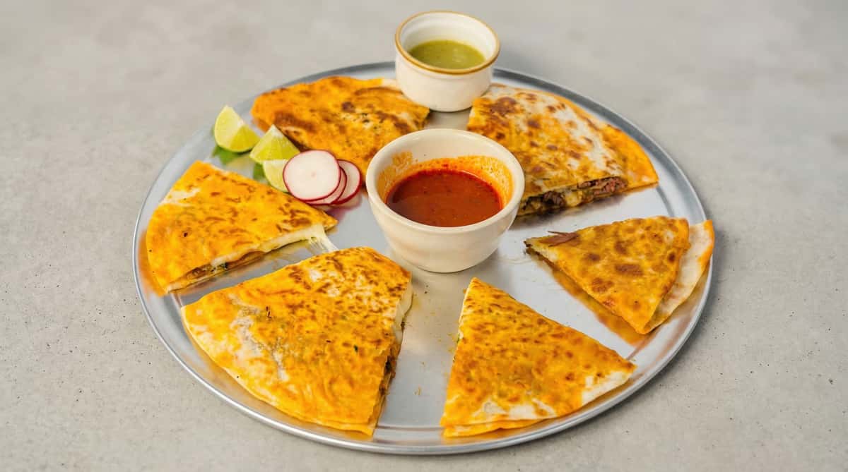 Cheesy beef quesadilla slices arranged on a platter with red and green salsa, lime wedges, and radishes for dipping