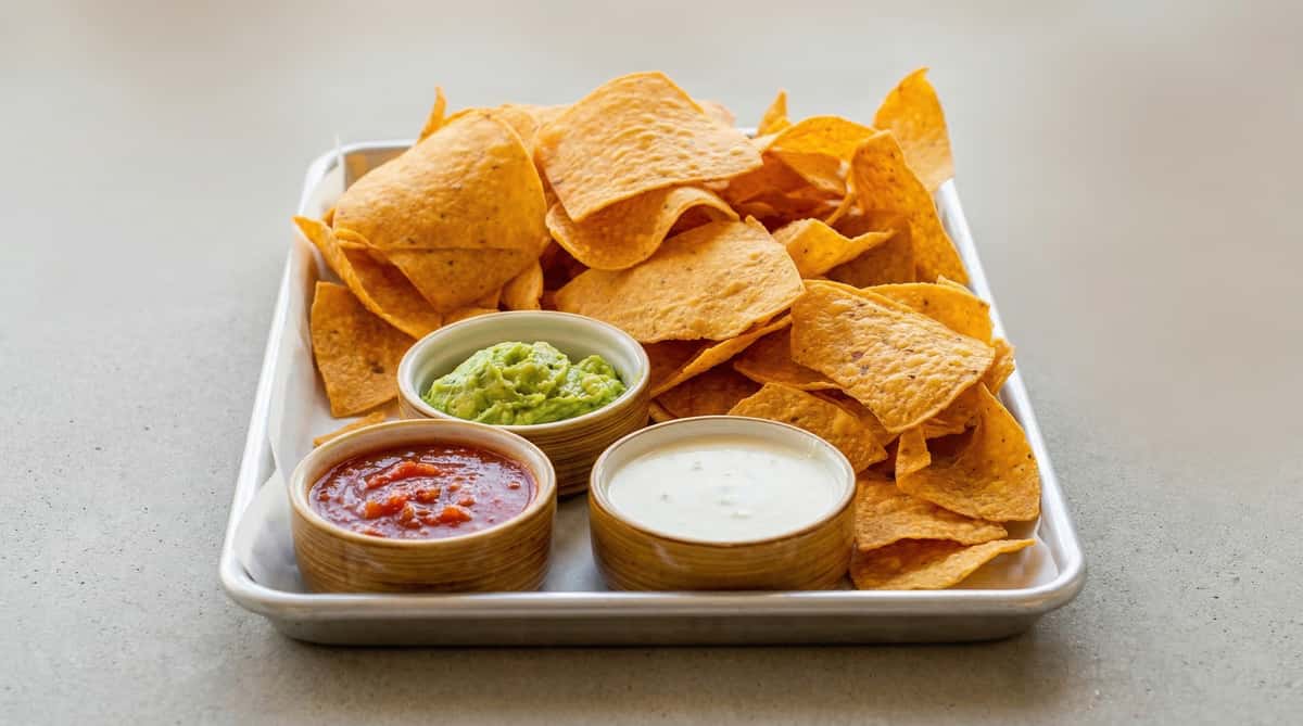 Crispy tortilla chips served with trio of dips including guacamole, salsa roja, and creamy queso dip on a metal tray