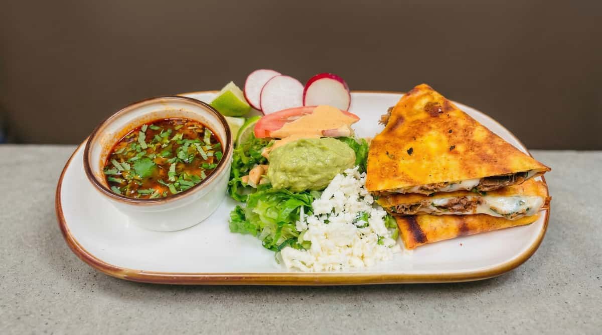 Grilled quesadilla with melted cheese and shredded meat, served with consommé, guacamole, salad, and queso fresco