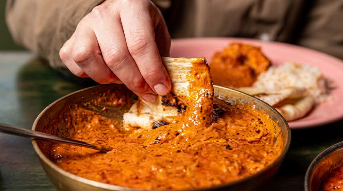 Fresh naan bread being dipped into a rich, creamy Indian curry surrounded by traditional accompaniments.