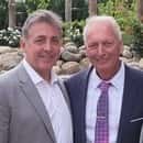 Two men in suits smiling outdoors, one in a gray jacket and white shirt, the other in a navy suit with a pink patterned tie.
