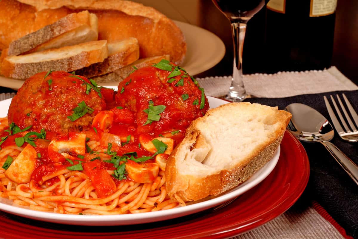 A hearty plate of spaghetti topped with rich tomato sauce, large meatballs, and chopped herbs, served alongside a slice of crusty bread and a glass of red wine. Perfect for a cozy Italian dinner.