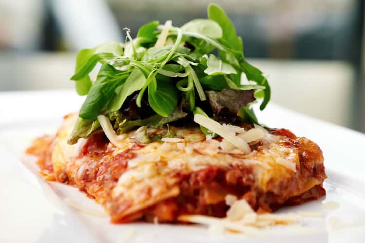 Classic lasagna topped with melted cheese, fresh arugula, and shaved parmesan on a white plate.