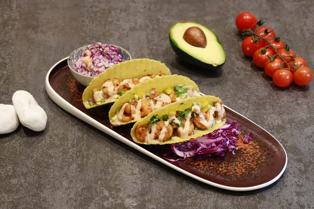 3 shrimp tacos and coleslaw on a tray with avocado and cherry tomatoes on the side