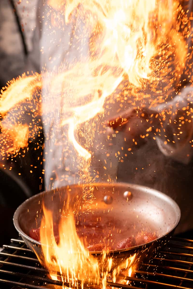 Chef flambés meat in a hot pan with tall flames and sparks rising dramatically in a professional kitchen setting.