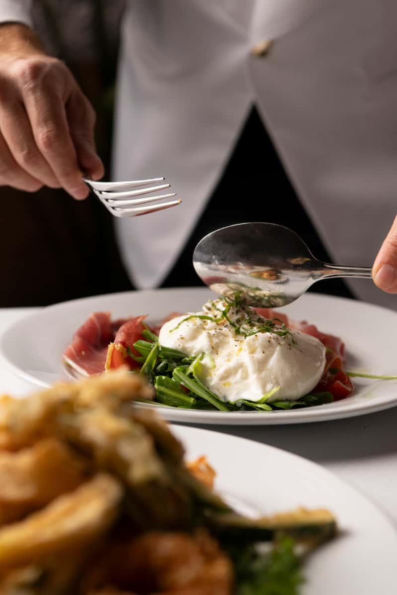 Burrata served on a bed of arugula and prosciutto, garnished with herbs and olive oil, as a person prepares to eat.