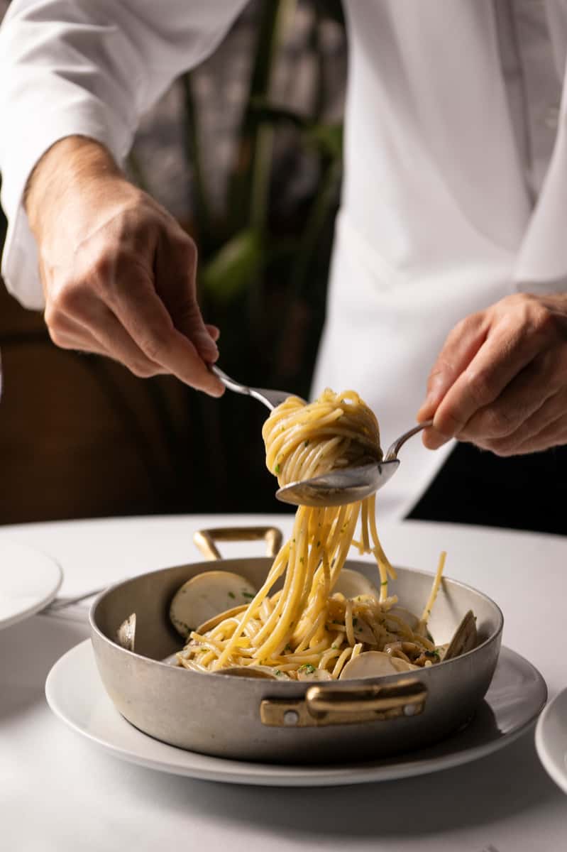 Chef plating linguine with clams in a creamy garlic sauce from a pan with golden handles on a white table.