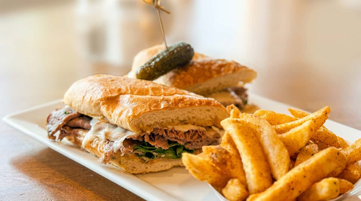 Italian beef sandwich on a baguette with melted cheese and lettuce, served with seasoned fries and a pickle.