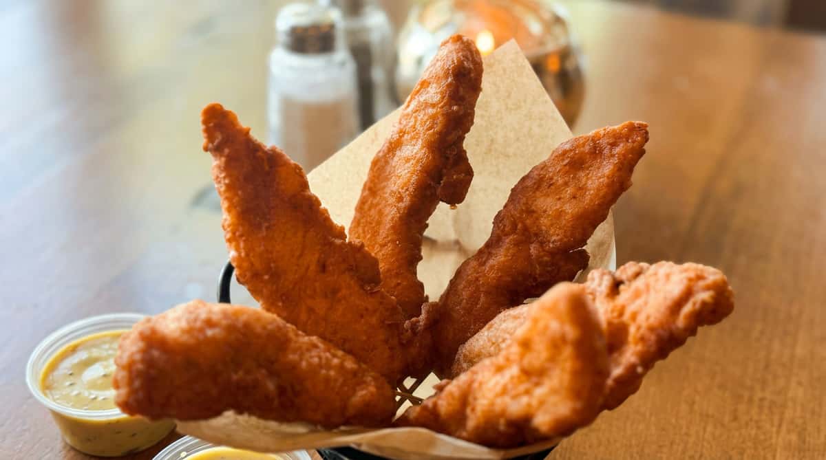 Fried battered chicken tenders in a black wire basket with parchment paper. A small container of honey mustard sauce is visible.