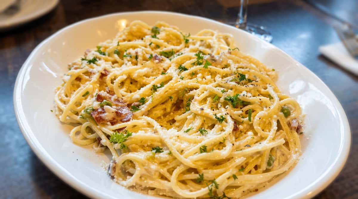 Spaghetti carbonara with creamy white sauce, bacon, green herbs, grated cheese, and black pepper.