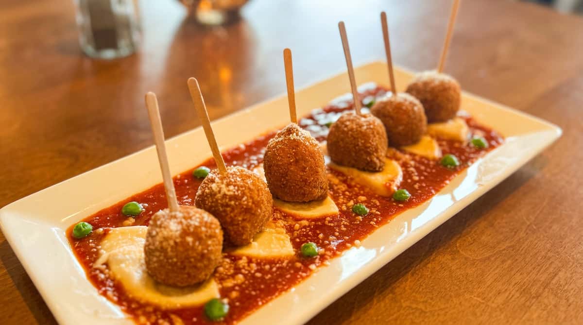 Five breaded arancini balls on skewers, atop pasta squares and red sauce, sprinkled with grated cheese and green peas.