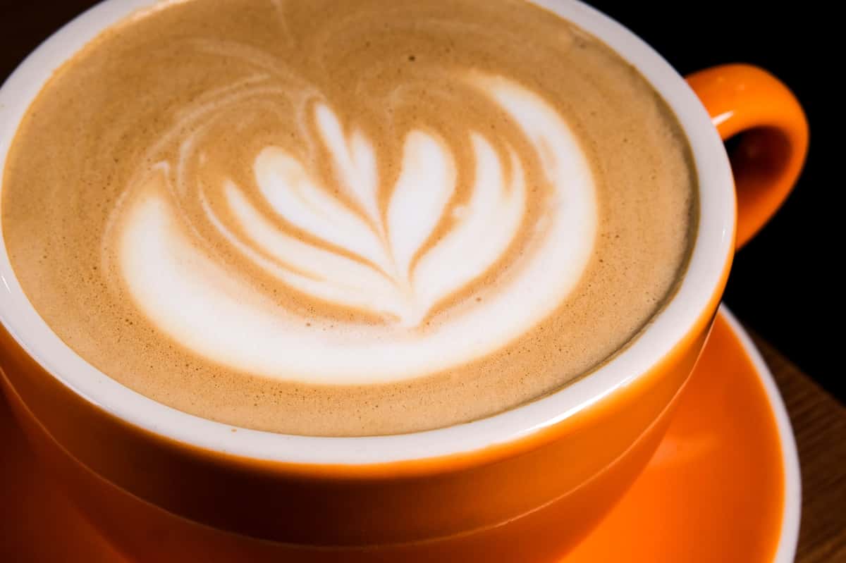 Latte with leaf-shaped latte art served in an orange cup and saucer on a wooden table in a cozy coffee shop.