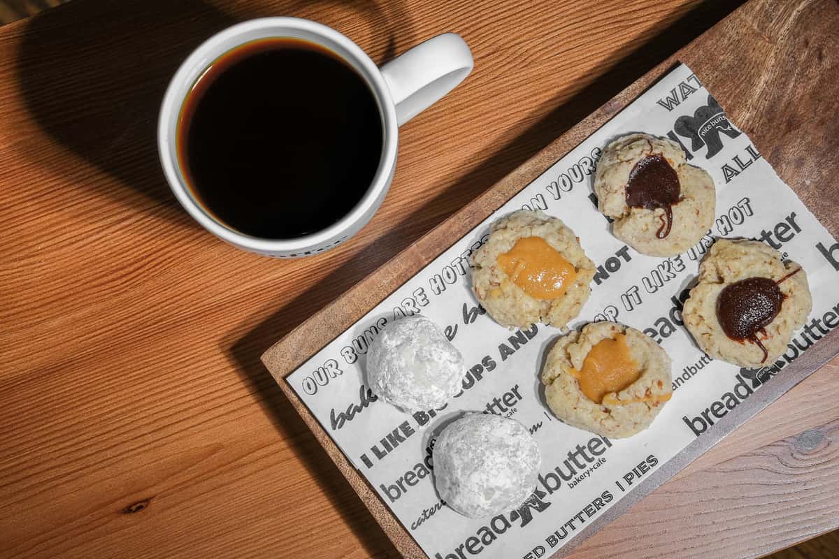 Tray of assorted cookies with powdered sugar and filled centers beside a white mug of black coffee on a wooden table.