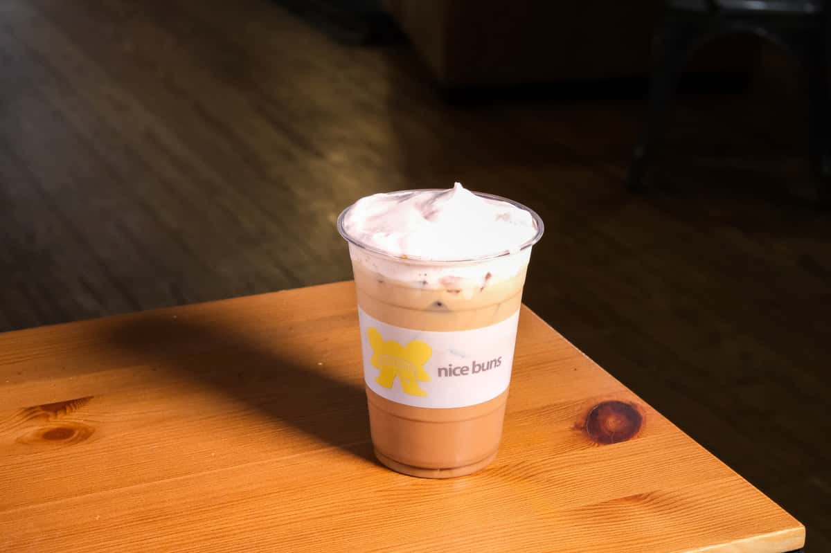Iced coffee topped with whipped cream in a clear plastic cup labeled "nice buns," placed on a wooden table.