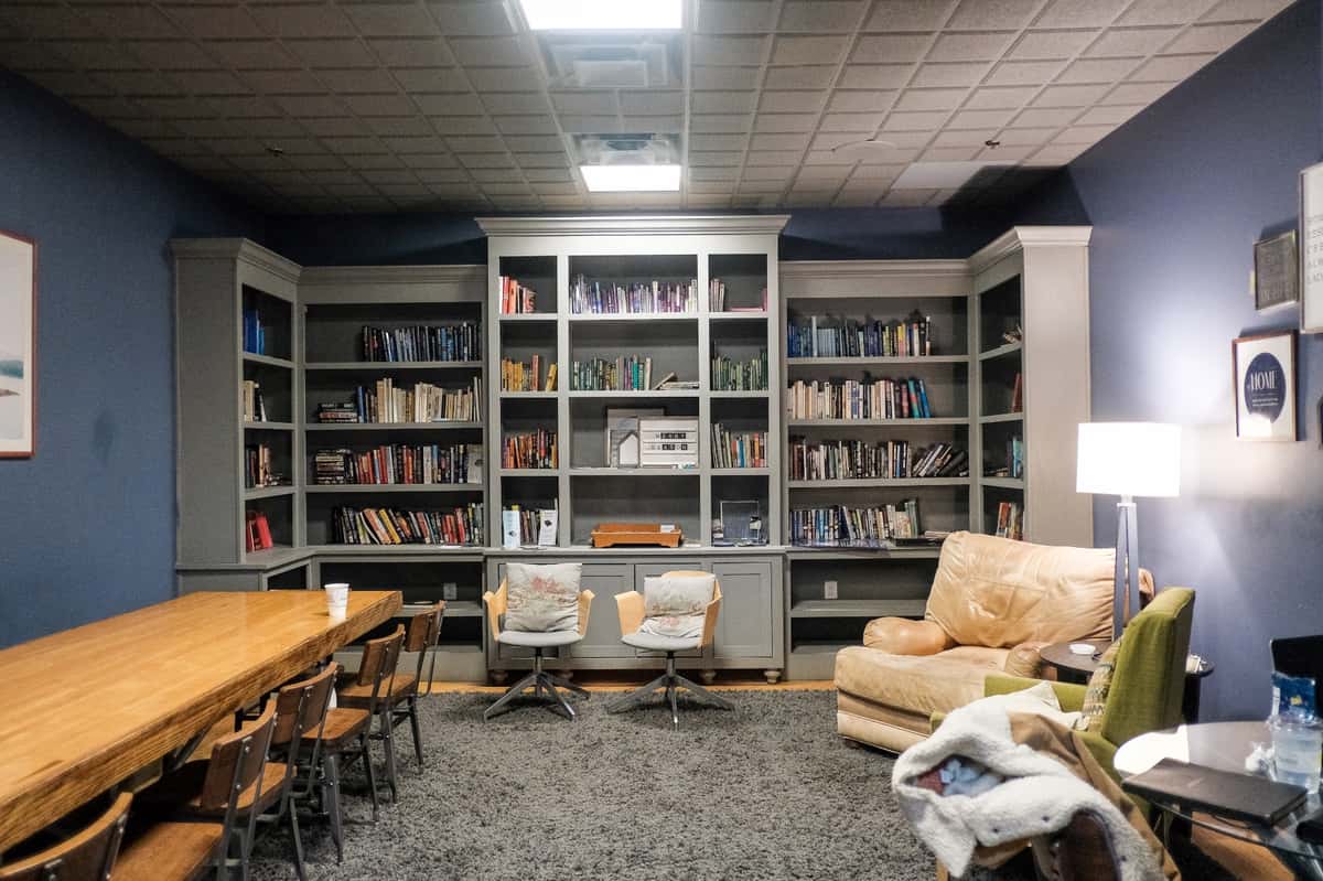 Cozy library-style room with built-in bookshelves, long wooden table, lounge chairs, sofa, and soft lighting for reading or small gatherings