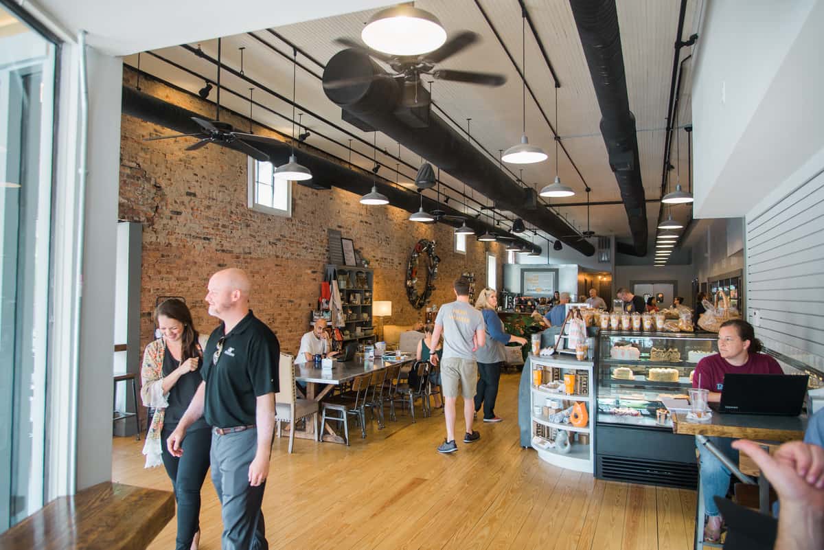 Bright, modern bakery café interior with exposed brick walls, communal seating, pastry display case, and guests enjoying coffee and freshly baked treats.