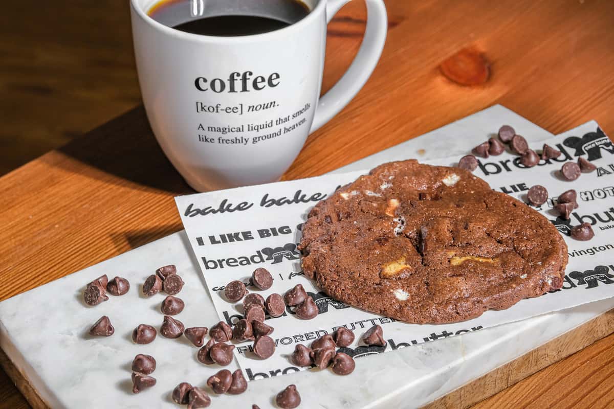 Freshly brewed coffee paired with a large double chocolate chip cookie at Bread & Butter Bakery and Cafe, served on a marble board with chocolate chips for a cozy café treat.