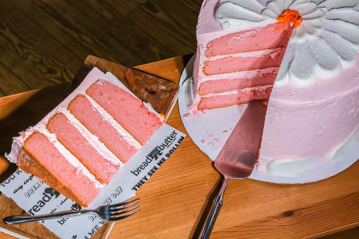 Pink layered cake with white frosting, one slice served on parchment with a fork and the remaining cake partially cut on a wooden table