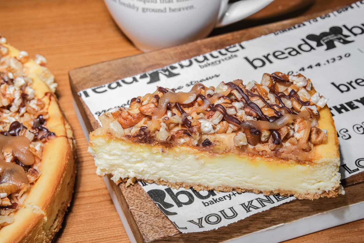 Slice of cheesecake topped with chopped pecans, chocolate drizzle, and caramel on Bread & Butter bakery paper.